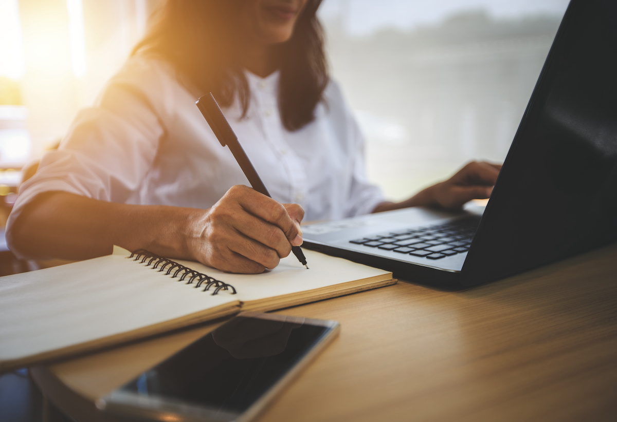 A young lady takes notes in a notebook while looking at a laptop.