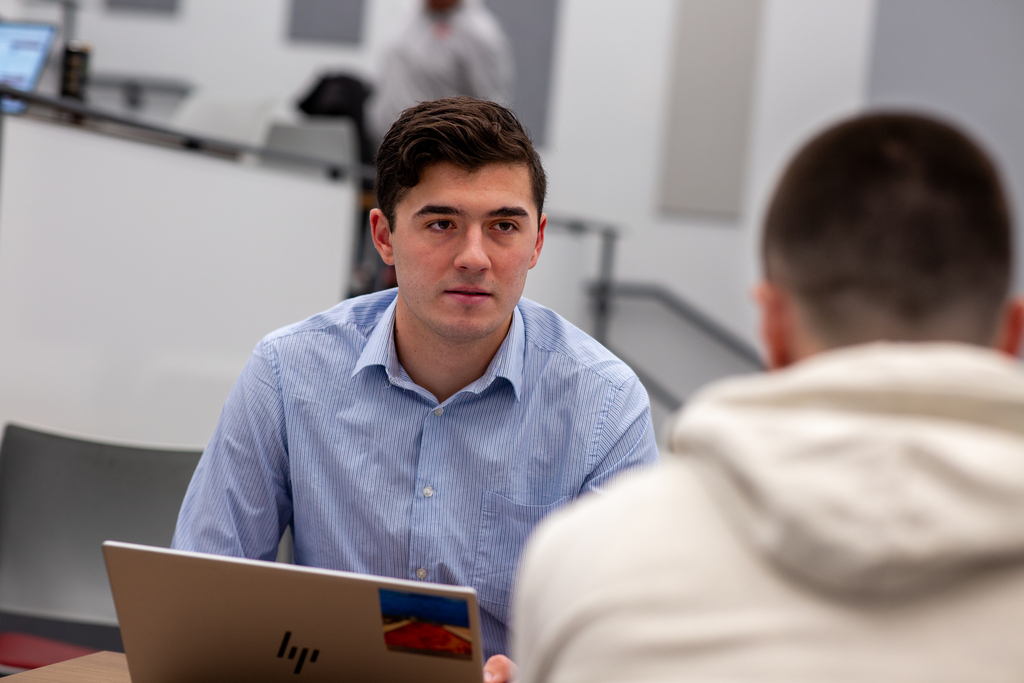 A young man sits behind a computer and talks to someone facing them.