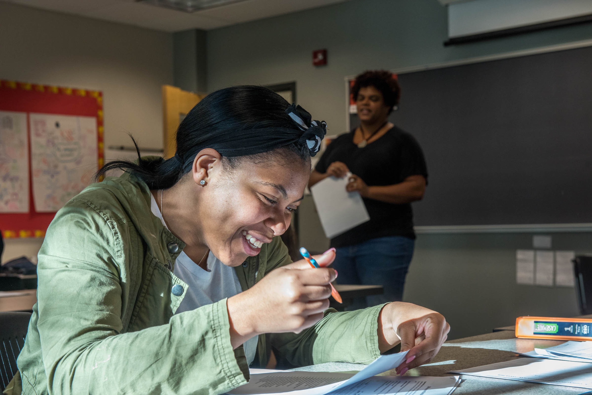 A young student smiles as a professor lectures in the background.