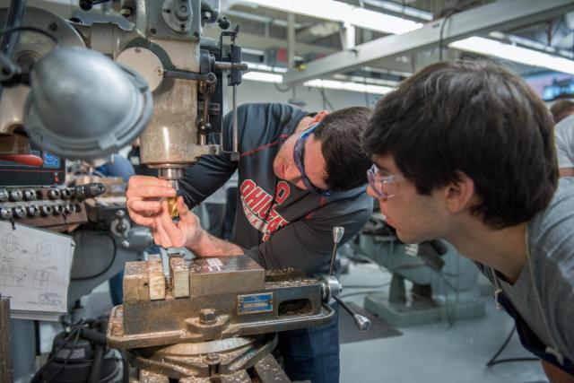 Two students examine a drill press in class.