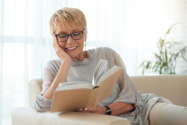 A smiling woman relaxed while reading a book on a sofa. 