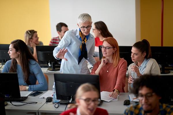 An instructor speaks to a couple students in a computer lab classroom.