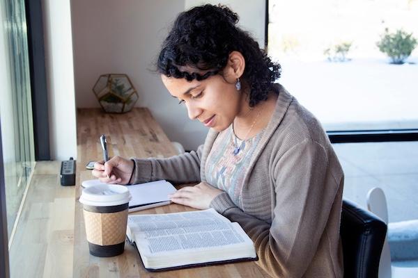 An instructor sits at a table reading with coffee, a book and a notepad.