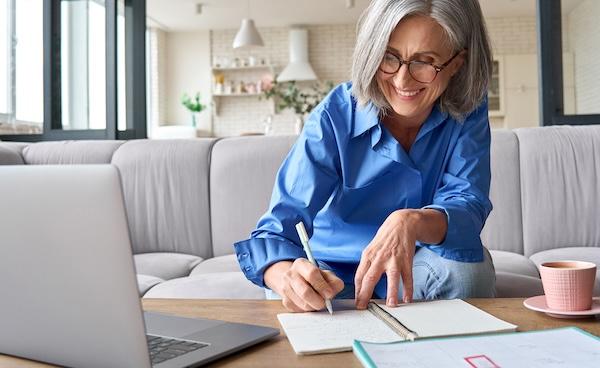 A smiling person with gray hair and glasses takes notes in front of an open laptop.