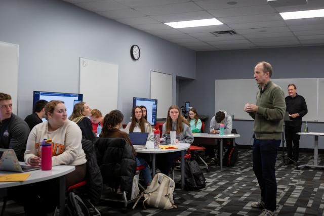 A professor stands in a classroom and instructs students seated at tables.