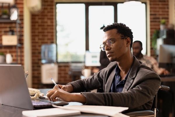 An instructor sits at a workspace in a common room reading on a laptop.