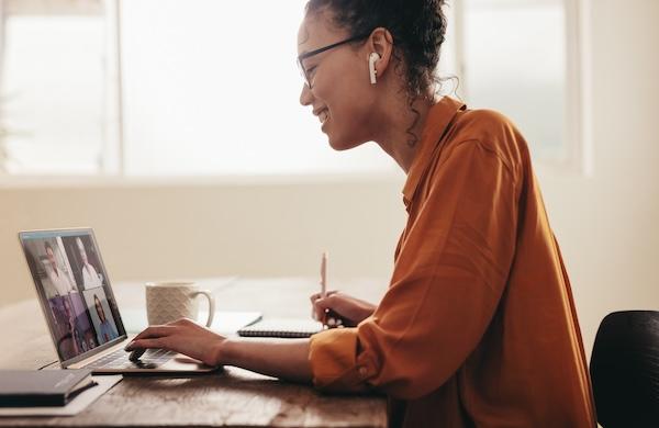 A smiling instructor wearing headphones talks with peers in a virtual workshop and takes notes.