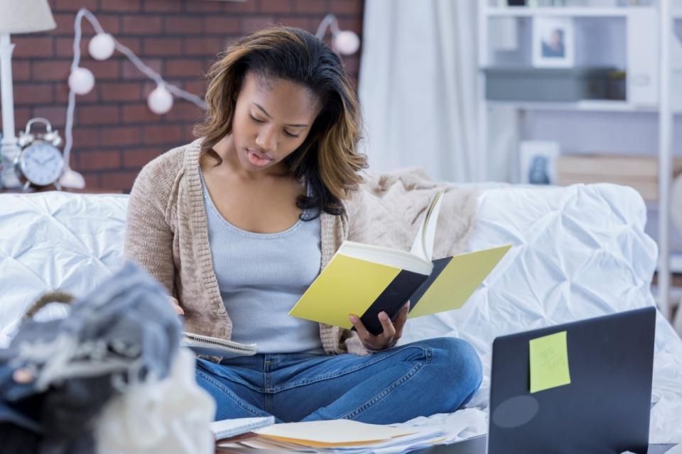 A student takes notes while reading a book with an open laptop in front of her.