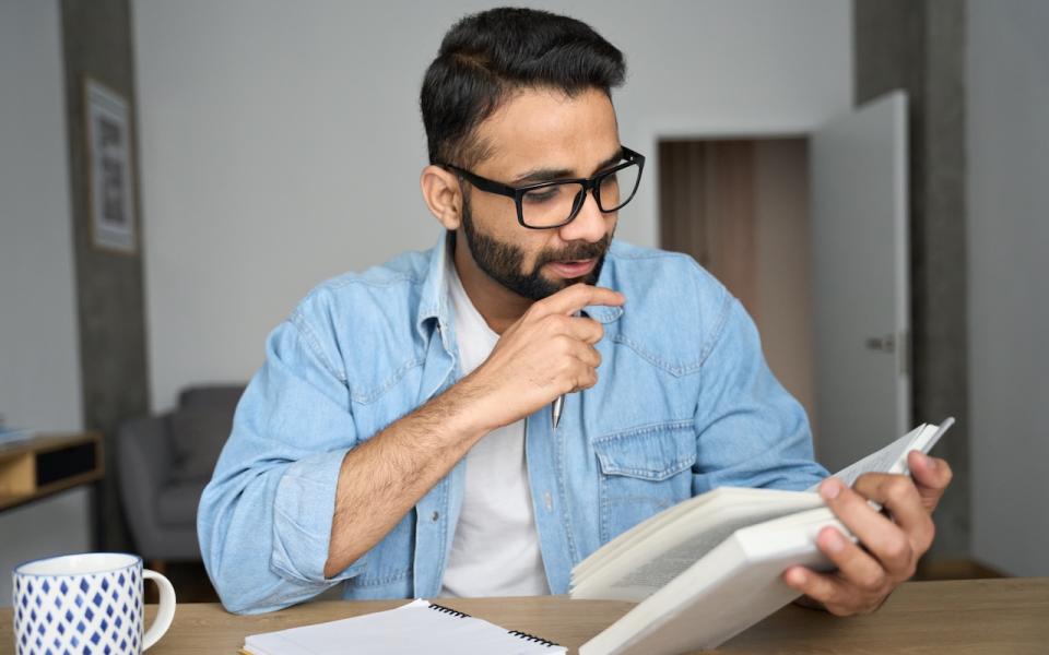 A man thoughtfully reading a book with a coffee and a notepad.