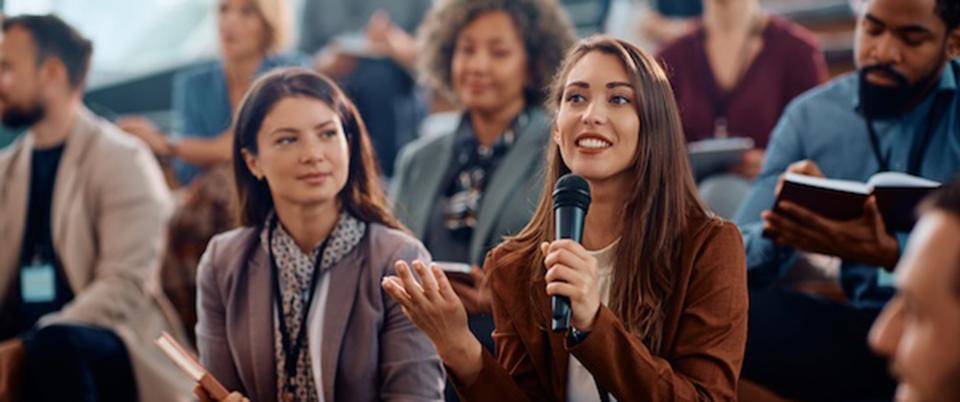 A woman holding a microphone asks a question at a conference session.