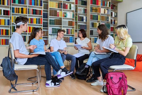 A group of students in discussion, sitting in a half circle against a wall of bookshelves.