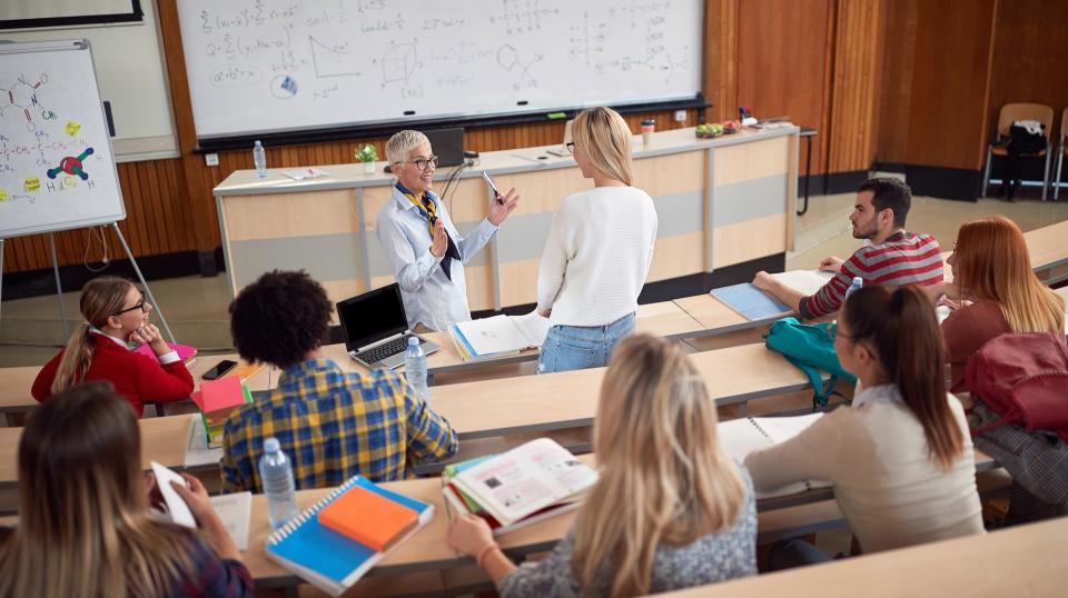 An instructor and one student talk at the front of a lecture hall while other students listen.